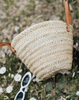 Woven straw bag with leather straps on a natural background with flowers and sunglasses.