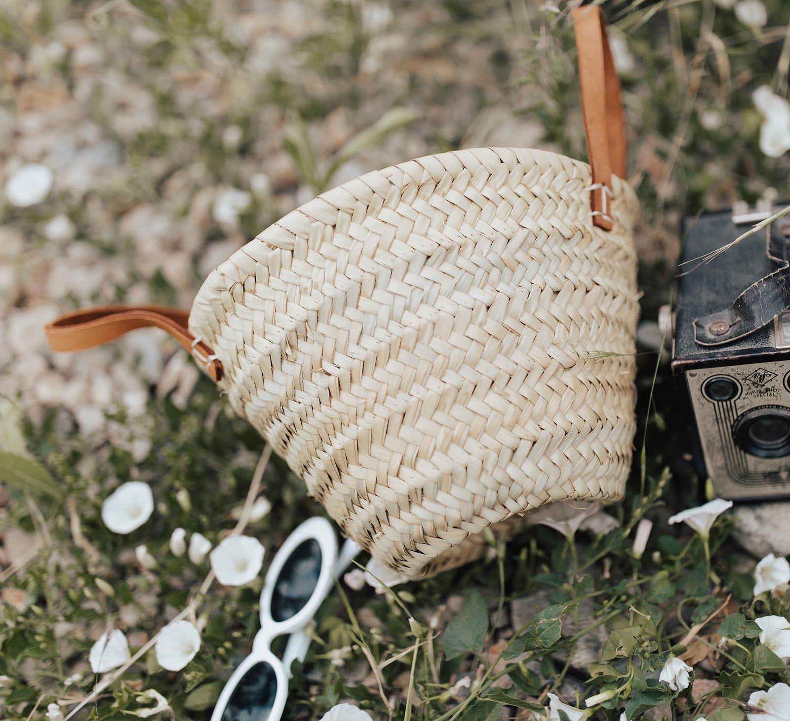 Woven straw bag with leather straps on a natural background with flowers and sunglasses.